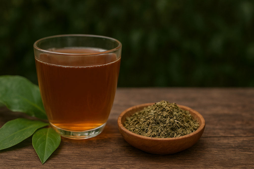 Sobre uma mesa de madeira uma xícara de vidro com chá ao lado de folhas verdes de cana do brejo e um pote de porcelana com folhas secas.