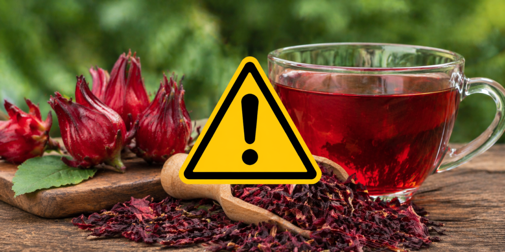 Sobre uma mesa de madeira uma xícara de vidro com chá e ao lado cálices frescos e secos de hibisco e por cima um símbolo de precaução para chamar atenção de quem não deve tomar chá de hibisco.