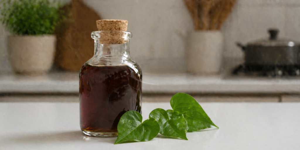 Em uma cozinha com destaque para um frasco de vidro com xarope de guaco e ao lado algumas folhas frescas de guaco sobre uma mesa branca. 